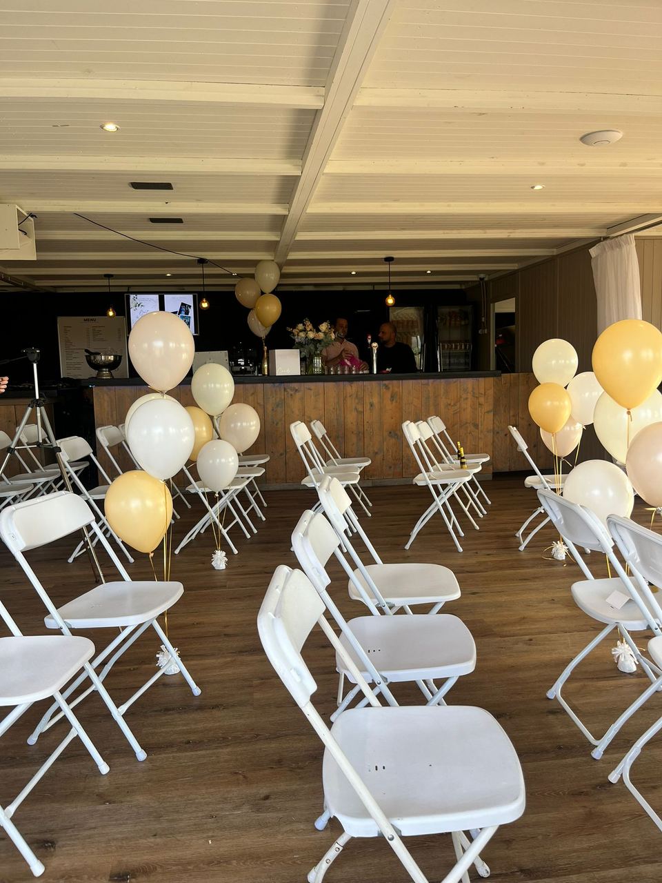 Zaal feestelijk ingericht met witte stoelen en gouden en witte ballonnen